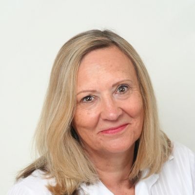 Head shot of a smiling Irene Kirkman who is wearing a white top and stood against a grey background looking directly into the camera